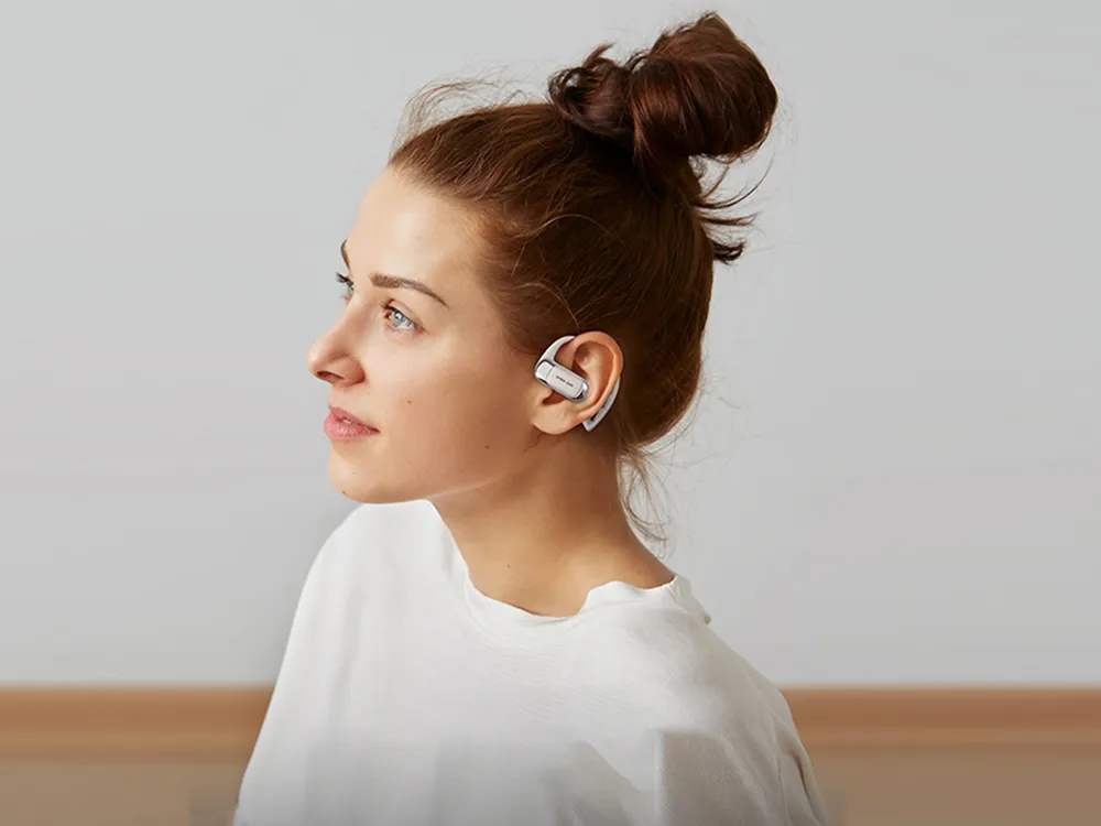Young woman with hair in a bun wearing Open Ear Buds that rest on her outer ear, allowing ambient sound awareness.