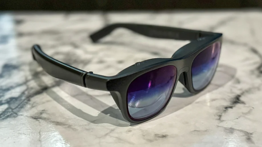 Black smart sunglasses with built-in electronics in chunky temples, reflective blue-purple lenses, resting on a marble surface.