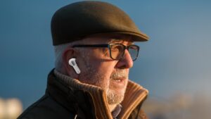 Close-up of an elderly man with glasses and a flat cap wearing white bone conduction hearing assist devices outdoors during sunset.