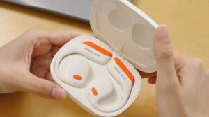 A close-up product shot of modern open-ear wireless headphones resting in their portable charging case. The earbuds feature a secure ear-hook design in a clean white finish with vibrant orange details and clear "OPEN EAR" branding, highlighting their comfortable, non-intrusive audio technology.