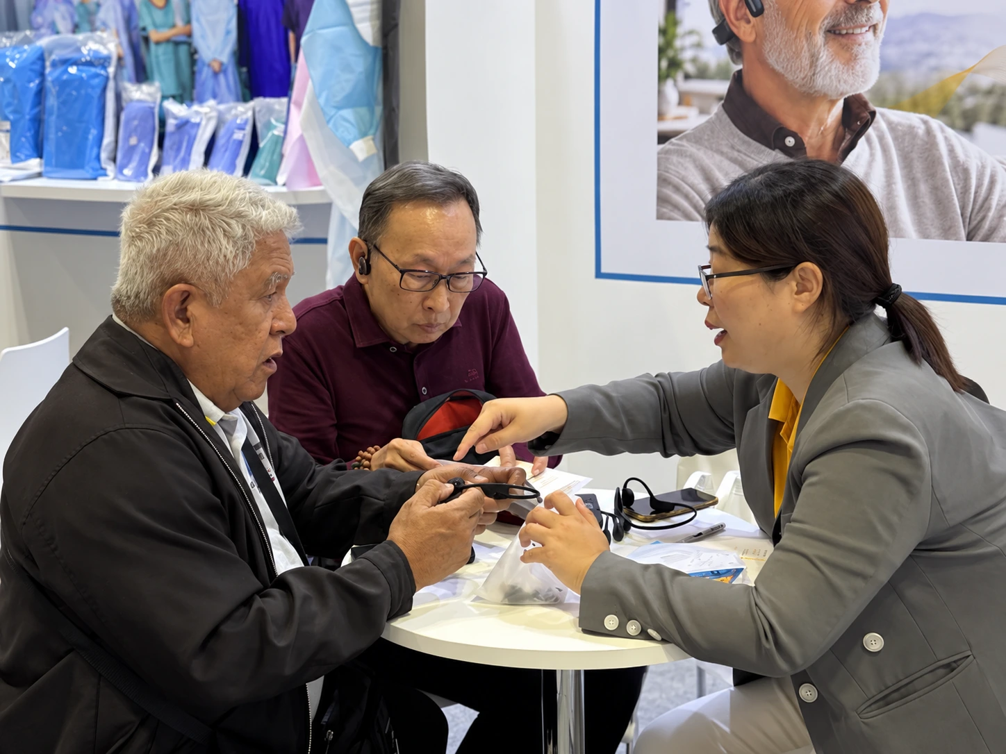Three people discussing an Alova hearing assistance device at an exhibition table